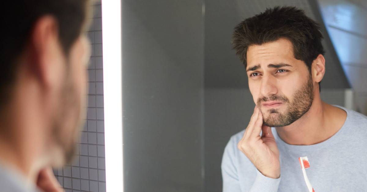 Hombre mirándose al espejo con un cepillo de dientes en una mano y la otra tocándose la cara como si tuviera un dolor de muelas. Esta imagen se utiliza para ilustrar una entrada sobre cómo aliviar un dolor de muelas.