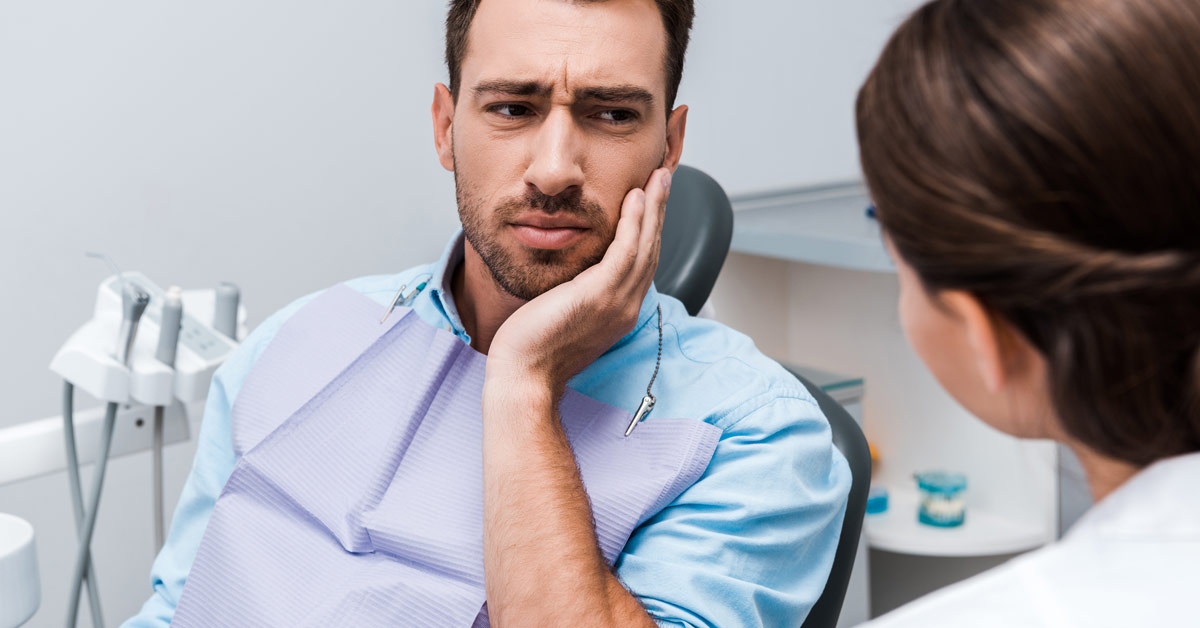 Hombre sentado en el sillón de la consulta del dentista con la mano tocando la mejilla, como indicando un dolor de muelas, mirando a la dentista que se encuentra frente a él.
