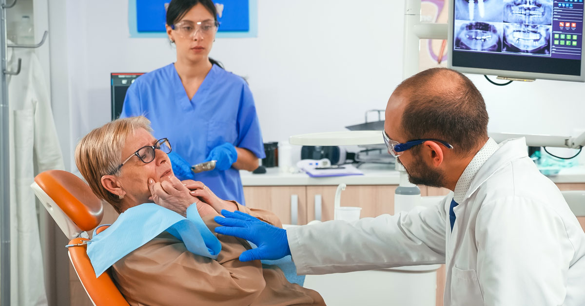 Mujer mayor señalando la mejilla al dentista en la consulta, como si explicara que esa es la zona de dolor. Esta imagen se utiliza para ilustrar una entrada sobre qué es la gingivitis.