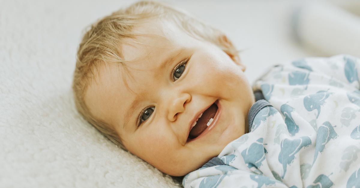 Primer plano de la cara de un bebé sonriendo acostado, mostrando sus primeros dientes. Esta imagen se utiliza para ilustrar una entrada sobre cuándo les salen los dientes a los bebés.